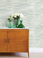 A wooden sideboard with ornate handles sits against a wall adorned with Lithos Marble Wallpaper (56 SqFt) by York Wallcoverings. On top of the sideboard are white flowers in green vases and small decorative glass bottles. The room boasts a light and minimalistic decor, enhancing its serene atmosphere.