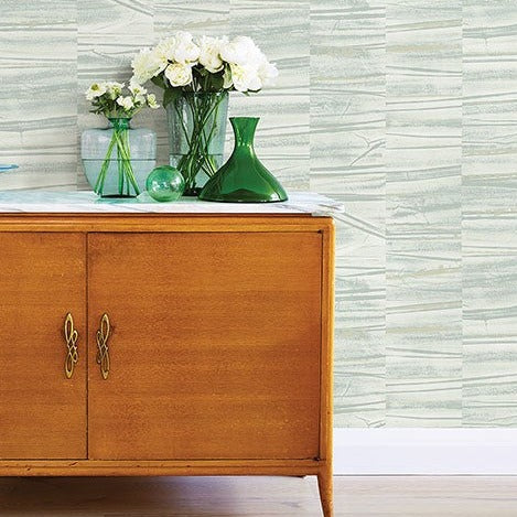 A wooden cabinet with brass handles stands against a wall adorned with Lithos Geometric Marble Wallpaper (56 SqFt) by York Wallcoverings. On top of the cabinet are two green glass vases, a clear glass ball, and a bouquet of white flowers in a glass vase. The floor is light wood.