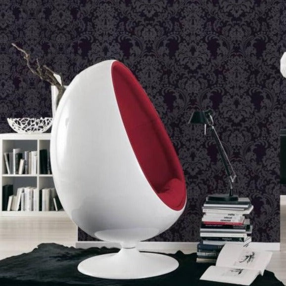 A modern white egg-shaped chair with a red interior is positioned on a black rug in a stylish room adorned with Ontario's Baroque Black Wallpaper. Nearby, there's a small stack of books, a black desk lamp, shelves filled with books, and a decorative white bowl.