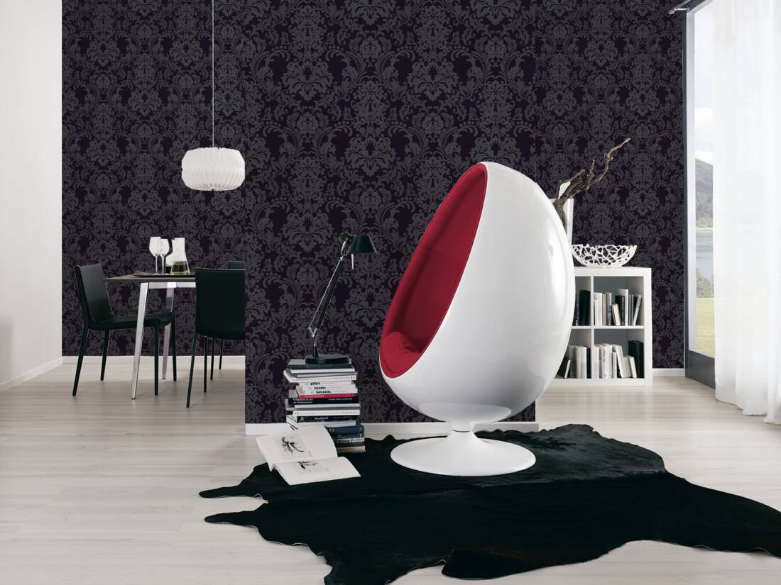 A modern living space featuring a black and white cowhide rug, a sleek white chair with a red interior, and a dark patterned accent wall. The small round dining table with two chairs sits under a pendant lamp. A stack of books is placed next to the chair as light floods through the window illuminating the Ontario Baroque White and Light Grey Wallpaper.