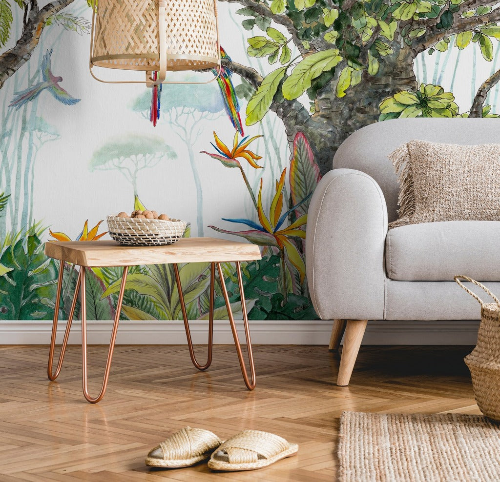 A cozy living room corner with a gray sofa, a wooden table with copper legs, vibrant Decor2Go Escape to the Jungle Wallpaper Mural featuring birds and foliage, a wicker lamp overhead, and slippers on the floor.