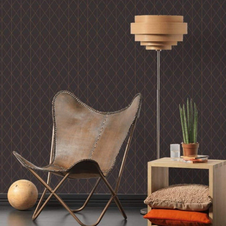 A modern, cozy interior features a brown butterfly lounge chair with a stitched design, a wooden geometric lamp, and a small wooden side table. The table holds a potted plant, an orange candle, and stacked cushions. The backdrop is an Ontario Diamond Geometric (56 SqFt) wallpaper with a diamond-patterned drop match.