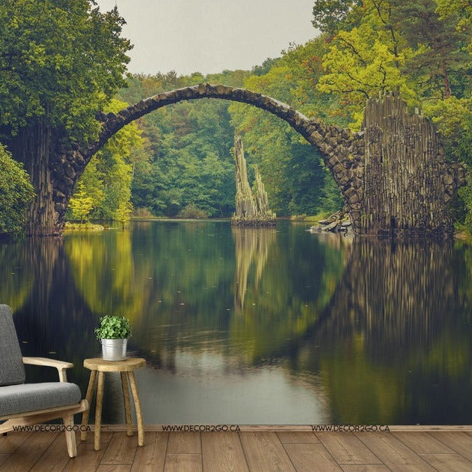 A serene indoor setting blending into a natural scene, featuring a modern armchair and side table set against a Decor2Go Wallpaper Mural depicting Circling Nature with a distinctive stone arch bridge surrounded by lush greenery.