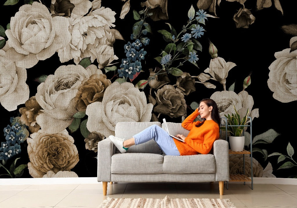 A woman in an orange sweater and jeans relaxing on a gray sofa, reading a book, against a dramatic backdrop of large Decor2Go Wallpaper Mural featuring peonies and blue flowers.