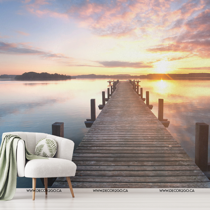 A tranquil sunset at a lakeside with a long wooden pier stretching into the water, encapsulating the essence of **Decor2Go Wallpaper Mural**. A stylish chair with a throw and a cushion, alongside a small table