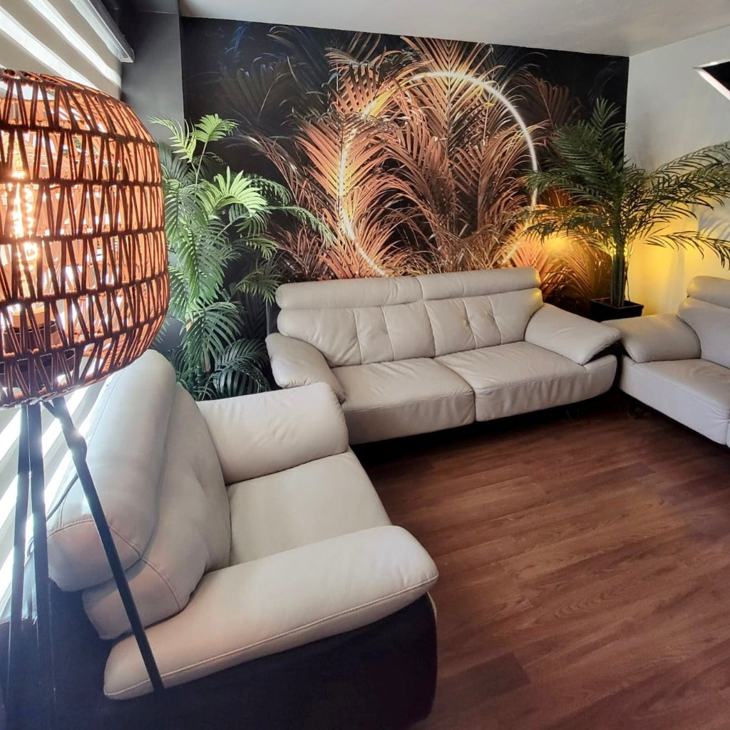 A modern living room featuring a large beige sofa against a bold "Big Green Leaves with Ring Light" wallpaper from Decor2Go Wallpaper Mural, with a tall, bamboo-style lamp casting a warm glow, and potted plants enhancing the room's ambiance.