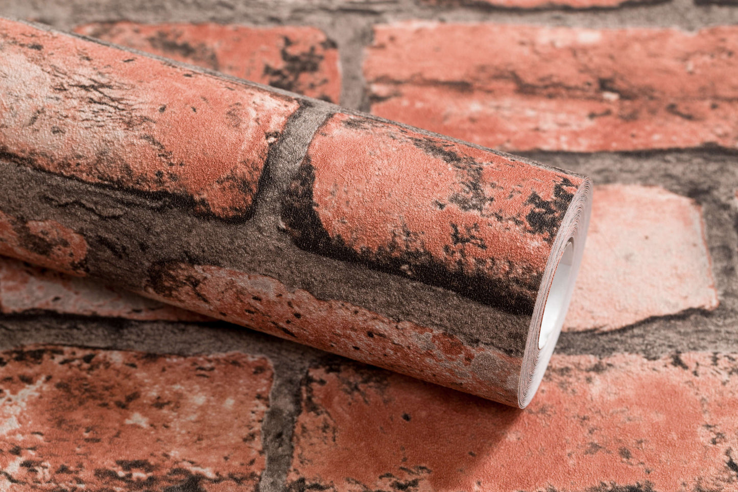 A close-up image of a roll of Palitra Brick Wall Wallpaper (114 SqFt) placed on a surface with a similar brick design. The wallpaper uses hot stamping to achieve a realistic texture that mimics the appearance of red bricks with dark mortar, enhancing the rustic look.