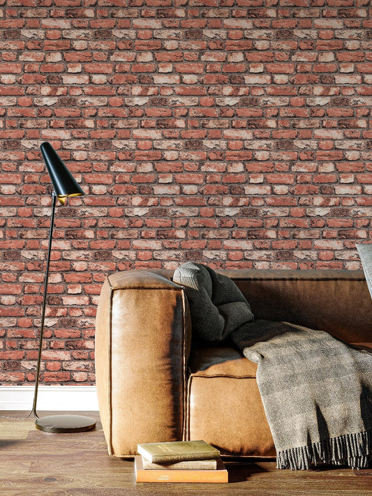 A cozy seating area with a light brown leather couch adorned with a gray throw blanket. Beside the couch is a modern black floor lamp. Books are stacked on the floor next to the couch. The backdrop features a wall covered with Palitra's Brick Wall Wallpaper (114 SqFt).