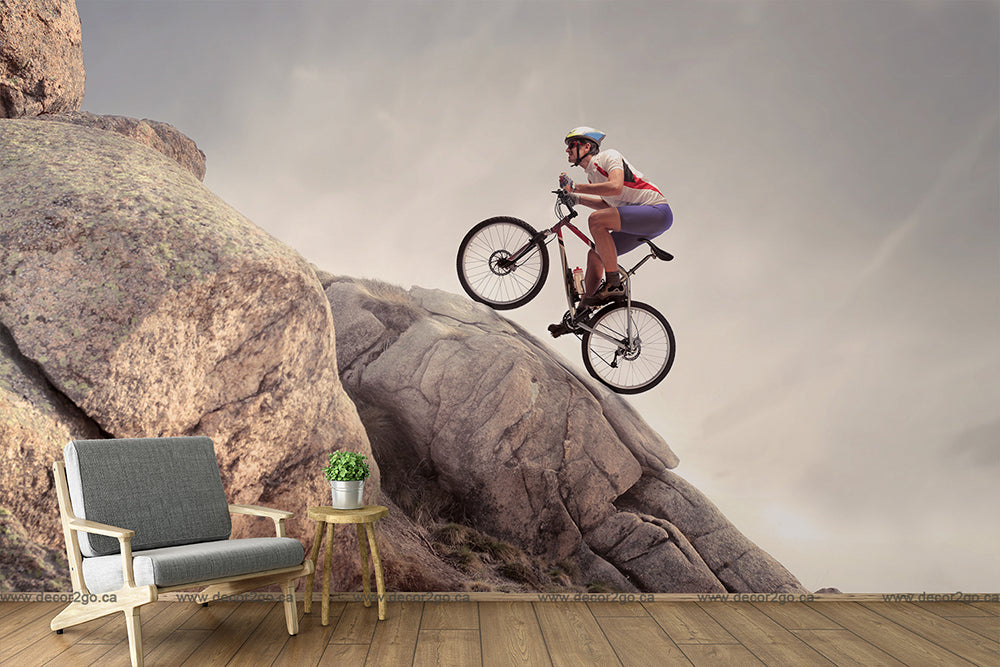 A surreal image blending indoor and outdoor elements, featuring a mountain biker in mid-air over a rocky outcrop with a modern armchair and a side table with a plant placed on a wooden floor can be found in the Decor2Go Wallpaper Mural.