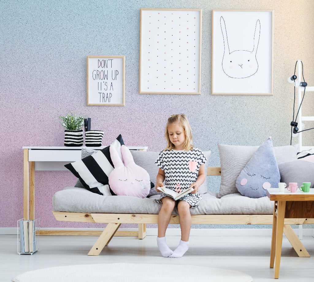 A young girl sits thoughtfully on a couch in a playfully decorated room with bunny-themed artwork and pillows, surrounded by soft colors and a cozy ambiance featuring Decor2Go Wallpaper Mural.