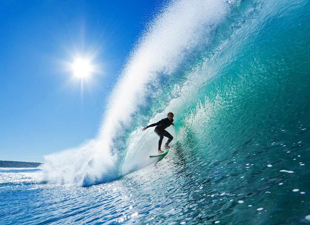 A surfer rides a large, clear blue wave with sunlight sparkling through it, highlighting the spray and intensity of the ocean in this Decor2Go Wallpaper Mural.