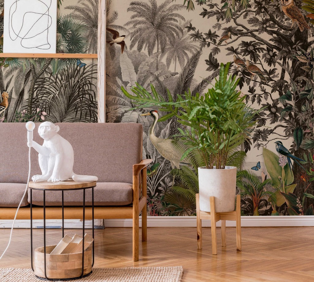 A stylish living room corner featuring a gray sofa, a small table with a white monkey figurine lamp, a wooden chair, a tall potted plant, and a wall covered in Decor2Go Wallpaper Mural's Vintage Jungle Wallpaper Mural.