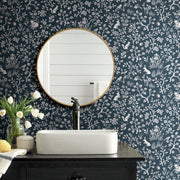 Bathroom - A features a round mirror with a gold frame, reflecting white shiplap walls. Below is a black faucet over a white rectangular sink on a dark wooden vanity. A vase of white tulips and a yellow sponge sit on the counter, which is adorned with York Wallcoverings' Fox & Hare (56 SqFt) prepasted wallpaper in a botanical pattern