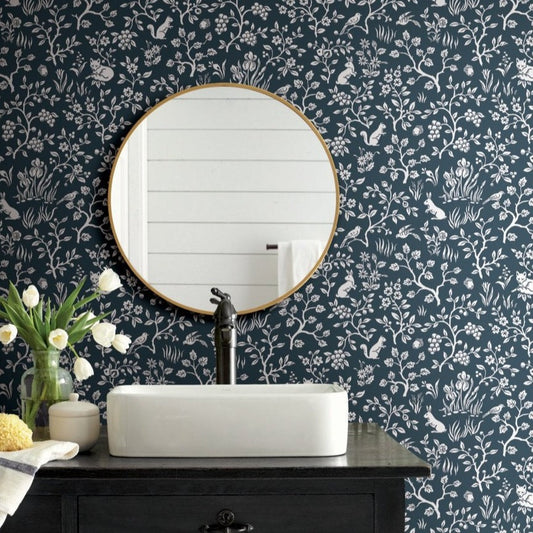 A bathroom features a round mirror with a gold frame, reflecting white shiplap walls. Below is a black faucet over a white rectangular sink on a dark wooden vanity. A vase of white tulips and a yellow sponge sit on the counter, which is adorned with York Wallcoverings' Fox & Hare (56 SqFt) prepasted wallpaper in a botanical pattern.