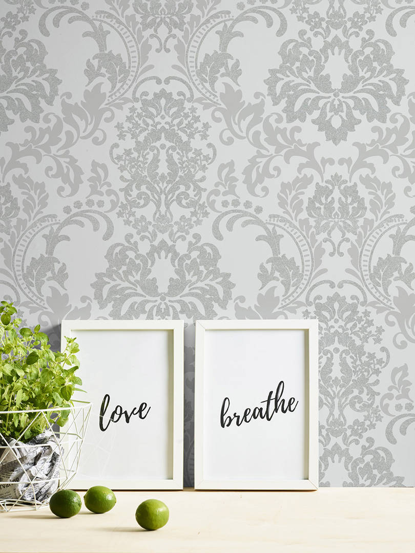 A Baroque Black Wallpaper by Ontario serves as the backdrop for a minimalist scene. Two framed prints with the words "love" and "breathe" lean against the wall. A wire basket with fresh green herbs and a few limes are placed on a light wooden surface.