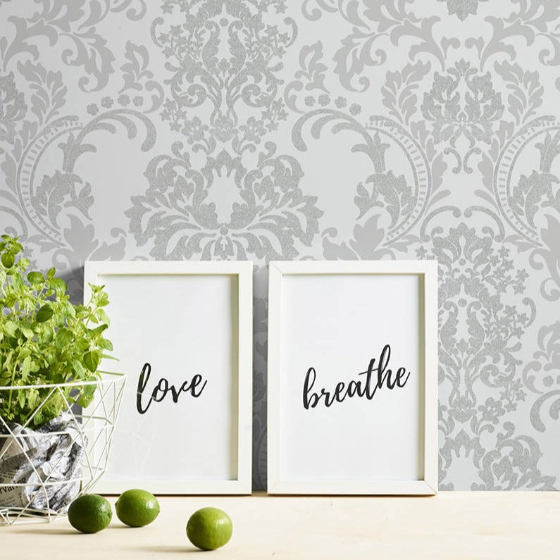 Two framed prints with the words "love" and "breathe" in cursive rest on a light wood textured surface with three green limes. Next to them, a white wire basket cradles a small green plant. The backdrop showcases elegant **Ontario Baroque White and Light Grey Wallpaper**.
