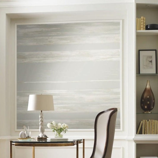A modern home office with a sleek white desk adorned with a white lamp, small vase of flowers, and stationery. Behind the desk is a large artwork featuring York Wallcoverings Horizontal Dry Brush Wallpaper (60 SqFt) in neutral tones. To the right, there are built-in shelves holding books, decor, and framed art.