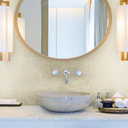 A modern bathroom features a round mirror with a wooden frame, two mounted lights on each side, and a stone basin sink with a wall-mounted faucet. The sink area includes a tray with toiletries and a white washcloth. The wall boasts York Wallcoverings' Geodes Layers Wallpaper (60 SqFt) for added sophistication.