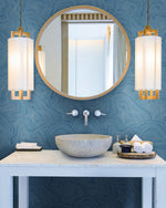 A modern bathroom features a round mirror above a white marble sink. Two cylindrical pendant lights with white shades and wooden accents flank the mirror. The walls boast a blue, textured pattern resembling Geodes Layers Wallpaper (60 SqFt) by York Wallcoverings. Accessories include towels, soap, and a small bowl on the counter.