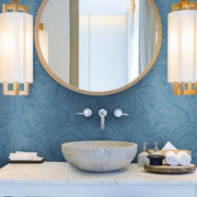 Bathroom - A modern features a round mirror with a wooden frame, mounted on York Wallcoverings' Geodes Wallpaper (60 Sq Ft) with a blue textured pattern. Below the mirror is a stone vessel sink with a wall-mounted faucet. Two contemporary pendant lights hang on either side. Various toiletries are arranged on the countertop