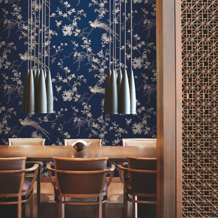 The dining room exudes elegance with a long wooden table and chairs. Modern pendant lights with a metallic finish hang above the table, while the walls are adorned with York Wallcoverings' Blue Bird And Blossom Chinoserie Wallpaper. A beautifully carved wooden partition adds a touch of sophistication on the right.