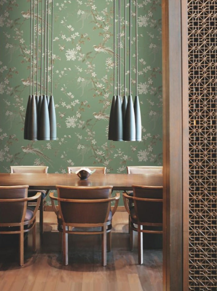 A dining area featuring a long wooden table and eight chairs is adorned with York Wallcoverings' Green Bird and Blossom Chinoiserie Wallpaper, showcasing beautiful Asian botanical motifs. Above the table, three clusters of modern pendant lights with a metallic finish are suspended, while an elegant touch is added on the right by a decorative wooden screen.