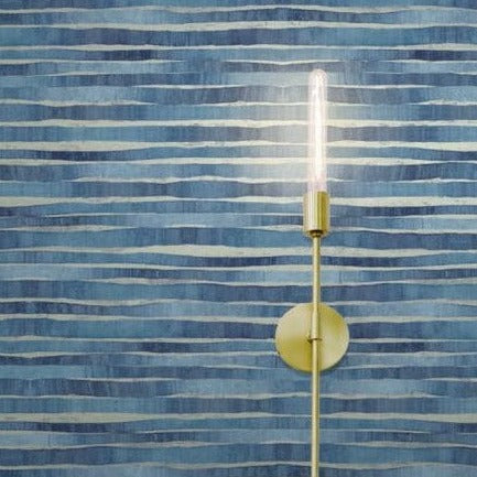 A minimalistic wall-mounted light fixture with a cylindrical, exposed bulb is positioned against Dreamscapes stripes (60 SqFt) by York Wallcoverings. The blue, striped wallpaper background features horizontal irregular lines in various shades of blue. The VinylCoated light fixture has a brass finish, adding to the mixed media landscape.