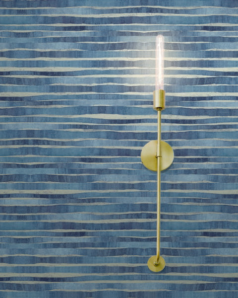 Gold wall-mounted light fixture with an exposed bulb extending vertically, set against York Wallcoverings' Dreamscapes Wallpaper (60 SqFt) featuring horizontal wavy lines in varying shades of blue. The metallic shimmer of the fixture and the illuminated bulb cast a warm glow, creating a dreamscape ambiance.