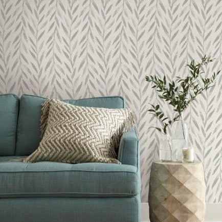 A cozy living space featuring a light blue couch with a chevron-patterned beige pillow. The wall behind the couch is adorned with Willow Wallpaper (56SqFt) by York Wallcoverings, vinyl-coated, leafy gray and white wallpaper. A geometric side table beside the couch holds a vase with green branches and a small candle.