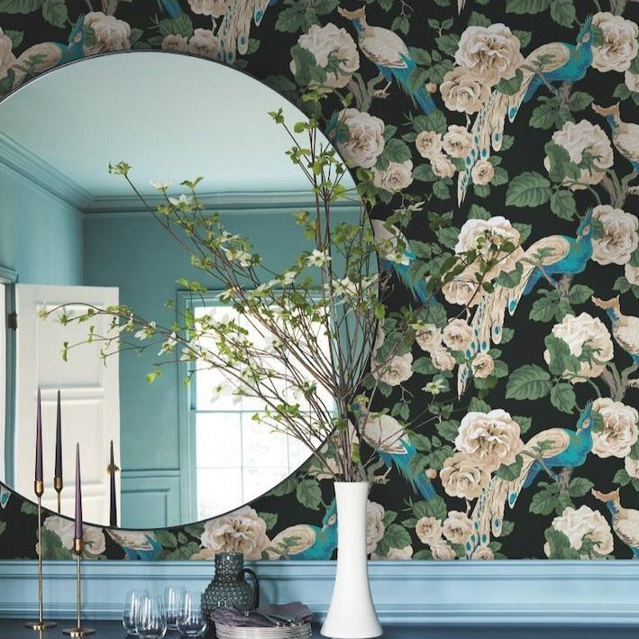 A circular mirror reflects part of a room adorned with York Wallcoverings' Garden Plume (60 Sqs.Ft), a green and beige floral and peacock-patterned wallpaper, showcasing the birds' colorful plumage. A white vase with a leafy branch sits on a table below the mirror. Three tall, thin candles and empty glasses are also on the table.