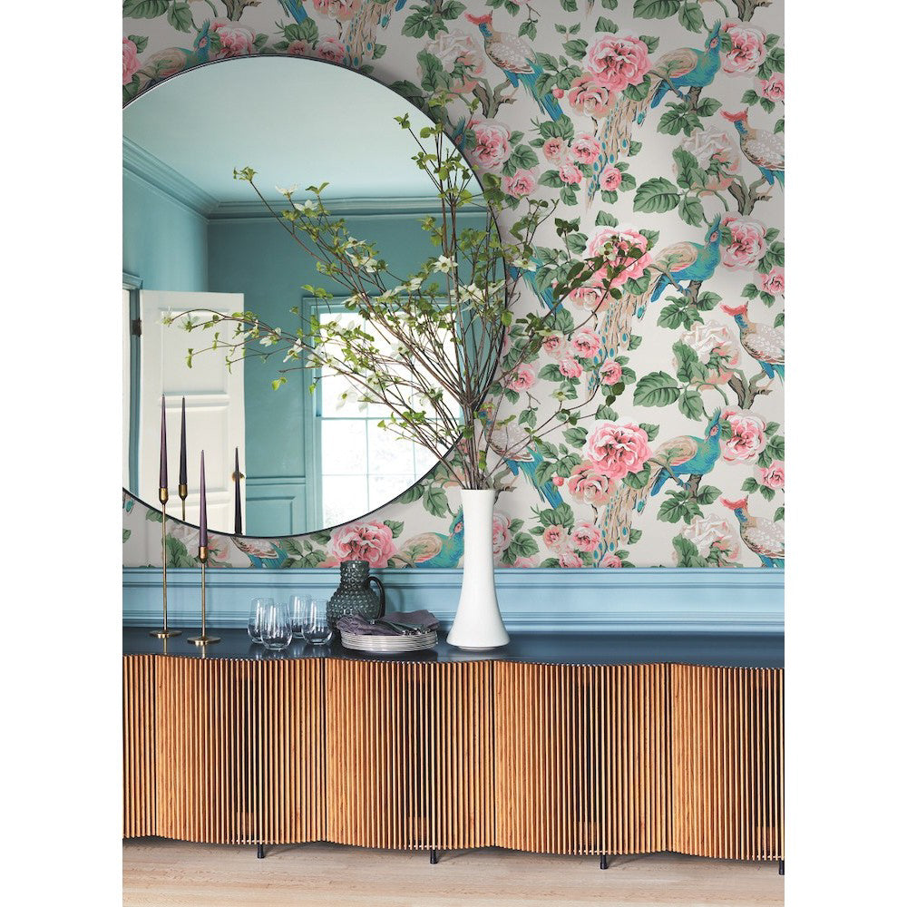 A stylish room features a large round mirror above a blue credenza with wooden slats. On the credenza, a white vase holds branches with small leaves. The peelable wallpaper behind is York Wallcoverings' Garden Plume (60 Sqs.Ft) with a floral and bird motif in pink, green, and blue tones. Four candle holders and glasses are also visible.