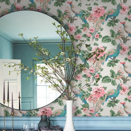 A round mirror reflecting a bright room with York Wallcoverings' Garden Plume Wallpaper (60 SqFt) featuring colorful plumage. A white vase with branches and green leaves is placed in front of the mirror. Three tall black candles and a few clear glasses are on a table nearby. The walls are painted teal.
