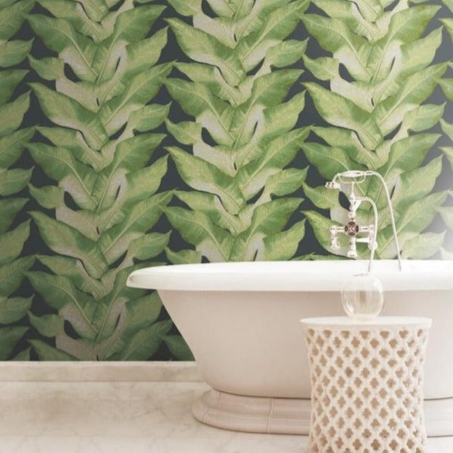 A bathroom with a modern aesthetic features a white freestanding bathtub and a matching stool. The focal point is the Beverly Hills Wallpaper (60 SqFt) by York Wallcoverings, adding a tropical touch with its bold, green leafy design. The floor is made of white marble, enhancing the Modern Heritage Design.