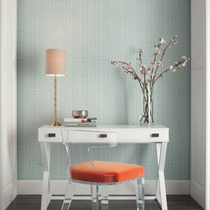 A modern study nook features a white desk with two drawers. A transparent chair with an orange cushion is placed in front. On the desk, there is a brass lamp with a beige shade, books, and a vase with pink cherry blossoms. The background has light blue Illusion Texture (60 SqFt) wallpaper by York Wallcoverings, enhancing the serene ambiance.