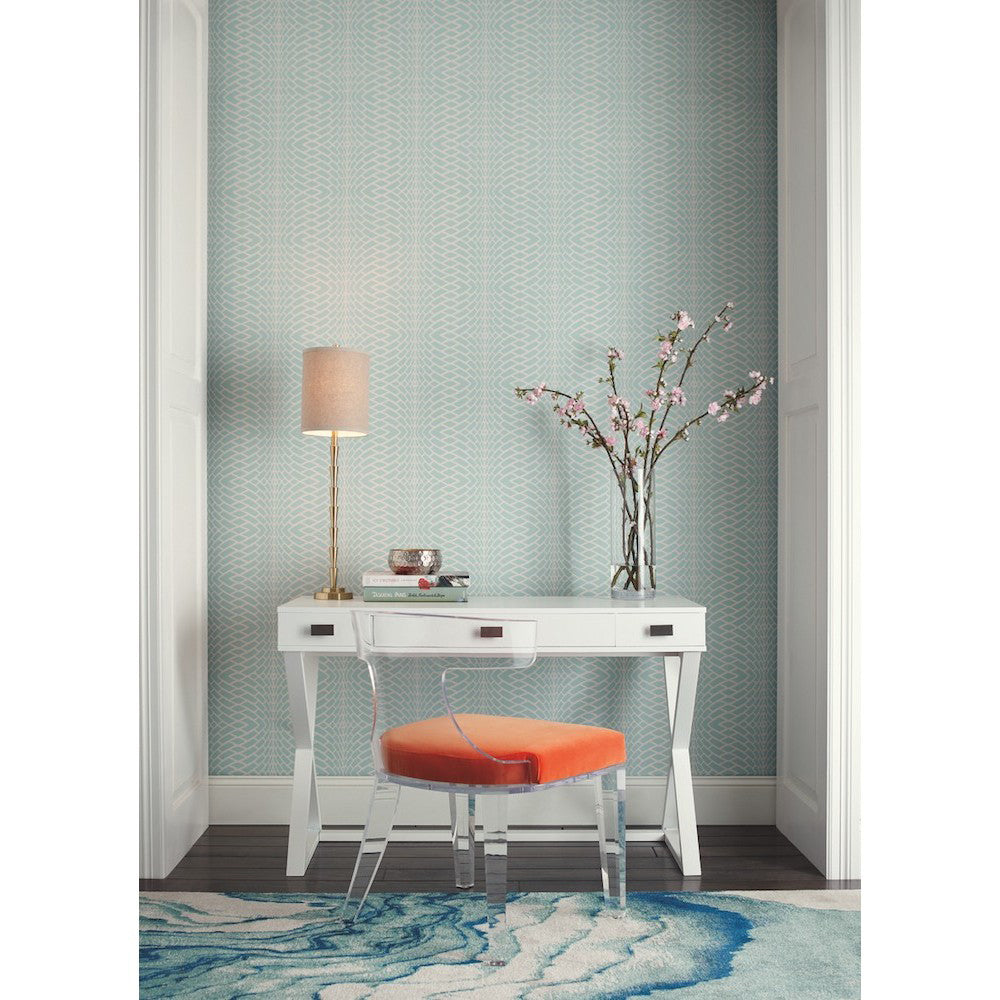 A modern workspace featuring a white desk with an orange cushioned chair. The desk holds a lamp, books, and a vase with pink flowers. The background showcases textured light blue Illusion Wallpaper (60 SqFt) from York Wallcoverings, and the floor displays a contemporary rug with wavy blue patterns.