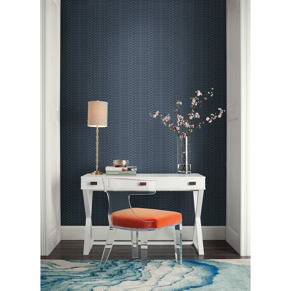 Home Office - A modern featuring a white desk with angled legs, a transparent chair with an orange cushion, a table lamp, and a vase with flowers. The room has York Wallcoverings Illusion Texture (60 SqFt) wallpaper in dark blue and a blue and white rug. A stack of books and a green box are on the desk