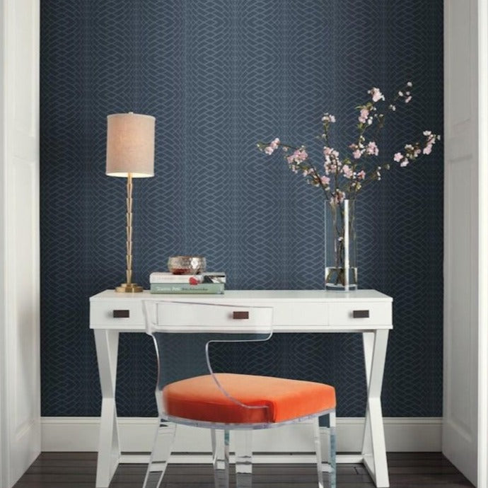 A modern, small workspace with a white desk and an orange cushion chair against a dark blue Illusion Wallpaper (60 SqFt) by York Wallcoverings geometric-patterned wall. On the desk are a brass table lamp, a glass vase with pink flowers, a stack of books, and decorative items. The floor is white wood.