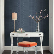 Home Office - A modern, small with a white desk and an orange cushion chair against a dark blue Illusion Wallpaper (60 SqFt) by York Wallcoverings geometric-patterned wall. On the desk are a brass table lamp, a glass vase with pink flowers, a stack of books, and decorative items. The floor is white wood