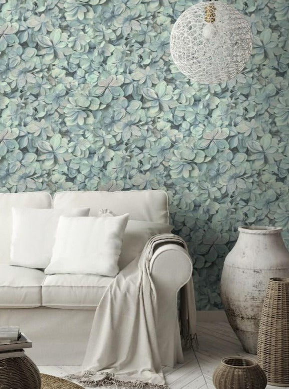 A cozy living room features a white sofa adorned with white cushions and a light beige throw blanket. The wall behind is beautifully covered with Hydrangea Wallpaper (56 SqFt) by York Wallcoverings, showcasing shades of green and blue. A large textured white pendant light hangs overhead, and a white ceramic vase sits nearby.