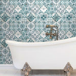 A white clawfoot bathtub stands against a wall adorned with intricate blue and white patterned tiles. The vintage-style brass faucet complements the bathtub's classic feet, enhanced by the use of York Wallcoverings' Mediterranean Tile Wallpaper, creating a luxurious and timeless bathroom setting.
