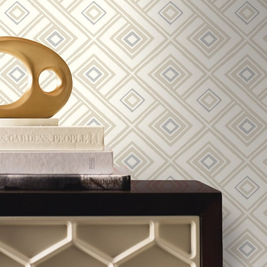 A decorative object with an abstract geometric design sits atop a small stack of books on a mantlepiece or shelf. The background features Pattern Play Paradox Wallpaper by York Wallcoverings, adorned with a diamond motif in shades of beige, cream, and gray, adding to the contemporary design.