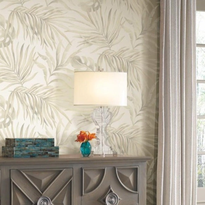 A gray wooden sideboard with geometric patterns holds a clear lamp with a white shade, an orange flower in a small blue vase, and a stack of decorative books. The background features Paradise Palm Leaves (60 SqFt) wallpaper by York Wallcoverings adorned with life-size leaves and a window with beige curtains.
