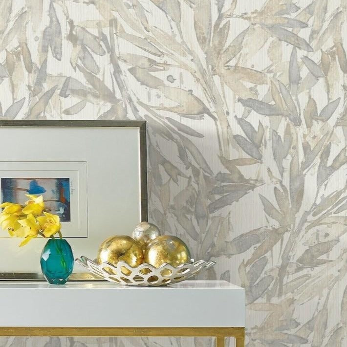 A white table with gold accents holds a small turquoise vase with yellow flowers, a framed artwork, and a decorative bowl containing golden spherical objects. The background showcases light, leaf-patterned York Wallcoverings Rainforest Leaves Wallpaper (56 SqFt) in neutral tones.