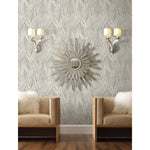 A stylish living room features a mirrored sunburst wall decor positioned between two modern, metallic wall sconces with white shades. Below, two beige armchairs with fluffy white pillows sit beside a striking Ebru Marble Texture (56 SqFt) by York Wallcoverings accent wall, and a wooden floor completes the cozy space.