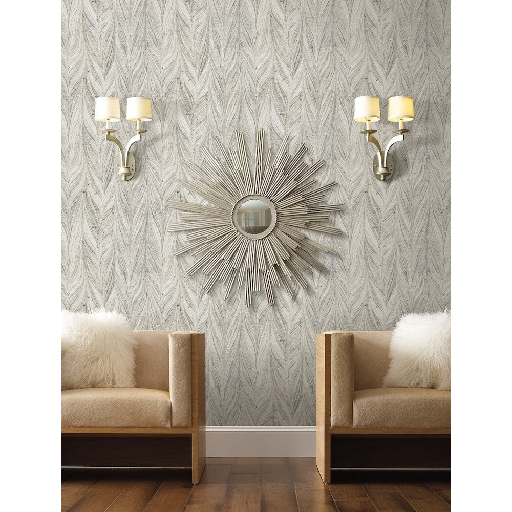 A stylish living room features a mirrored sunburst wall decor positioned between two modern, metallic wall sconces with white shades. Below, two beige armchairs with fluffy white pillows sit beside a striking Ebru Marble Texture (56 SqFt) by York Wallcoverings accent wall, and a wooden floor completes the cozy space.