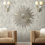 A modern sitting area features two beige armchairs with white fluffy pillows. Centered on the wall behind them, a round sunburst mirror is surrounded by silver rays. The background boasts York Wallcoverings' Ebru Marble (56 SqFt) luxury wallcoverings, while two elegant wall sconces with cream-colored shades flank the mirror, casting warm light.